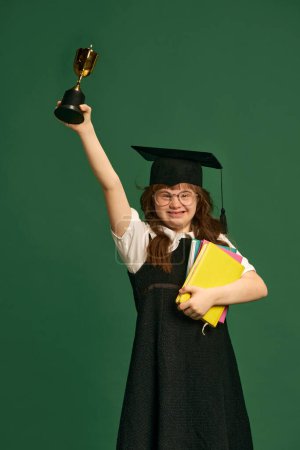 Téléchargez les photos : Fille heureuse avec le syndrome du duvet debout dans le chapeau de graduation, livres et trophée sur fond de studio vert. L'éducation. Concept d'acceptation, soins, inclusion, santé, diversité, émotions, égalité - en image libre de droit