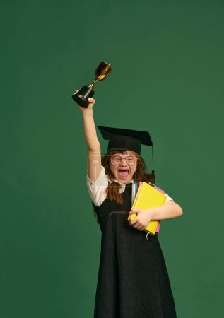 Photo for Happy girl with down syndrome standing in graduation cap, books and trophy against green studio background. Education. Concept of acceptance, care, inclusion, health, diversity, emotions, equality - Royalty Free Image