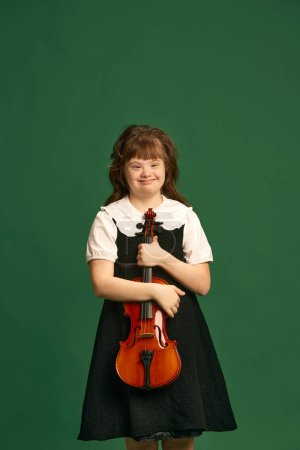 Téléchargez les photos : Belle fille avec le syndrome du duvet debout avec violon, apprendre la musique sur fond de studio vert. Concept d'acceptation, soins, inclusion, santé, diversité, émotions, égalité - en image libre de droit