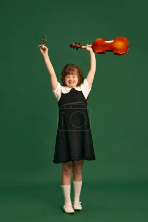 Photo for Beautiful girl with down syndrome standing with violin, learning music against green studio background. Concept of acceptance, care, inclusion, health, diversity, emotions, equality - Royalty Free Image