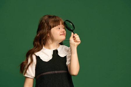 Photo for Teen girl with down syndrome looking in magnifying glass against green studio background. Education for people with special needs. Concept of acceptance, care, inclusion, health, diversity, equality - Royalty Free Image