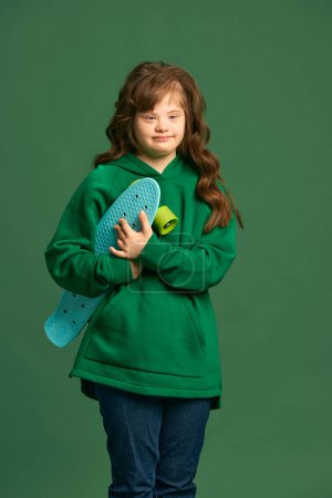 Photo for Teen girl with down syndrome wearing cozy hoodie, standing with skateboard against green studio background. Concept of acceptance, care, inclusion, health, diversity, emotions, equality - Royalty Free Image