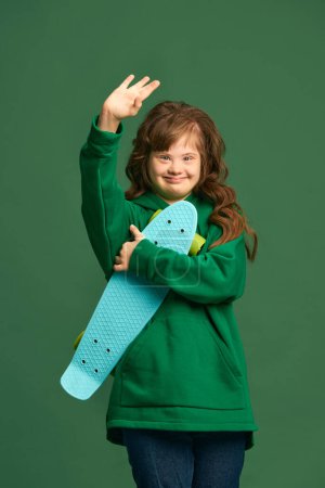Photo for Teen girl with down syndrome wearing cozy hoodie, standing with skateboard against green studio background. Concept of acceptance, care, inclusion, health, diversity, emotions, equality - Royalty Free Image