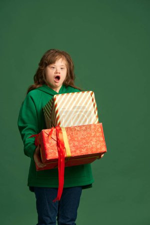 Photo for Emotional teen girl with down syndrome standing with socked face, holding many present boxes on green studio background. Concept of acceptance, care, inclusion, health, diversity, emotions, equality - Royalty Free Image