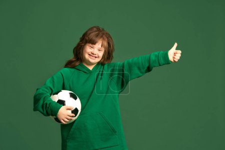 Photo for Happy smiling teen girl with down syndrome standing with football ball against green studio background. Sportive life. Concept of acceptance, care, inclusion, health, diversity, emotions, equality - Royalty Free Image