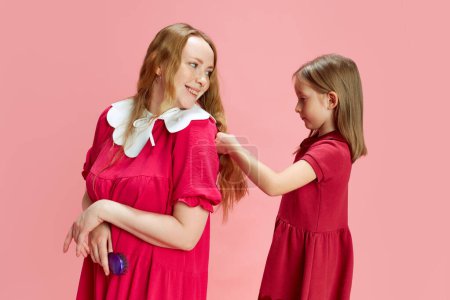 Photo for Like mom, like daughter. Little girl doing hairstyle to her mother against pink studio background. Family look. Concept of Mothers Day, International Happiness Day, fashion, childhood, care and love - Royalty Free Image