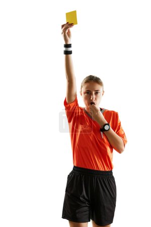 Photo for Young serious woman, soccer referee blowing a whistle and showing yellow card as warning symbol against white studio background. Concept of sport, competition, match, profession, control - Royalty Free Image