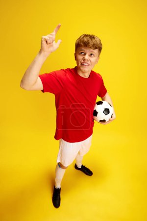 Foto de Retrato de vista superior de un joven con pecas en uniforme de fútbol, sosteniendo pelota de fútbol sobre fondo amarillo del estudio. Aficionado al deporte, apostando. Concepto de estilo de vida activo, juventud, afición y emociones humanas - Imagen libre de derechos
