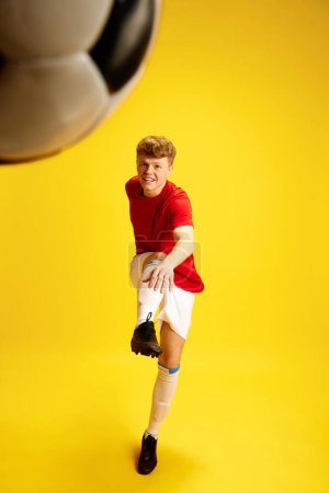 Photo for Dynamic image of young man in soccer sportswear, uniform training, hitting ball against yellow studio background. Concept of active lifestyle, youth, hobby and human emotions, game - Royalty Free Image