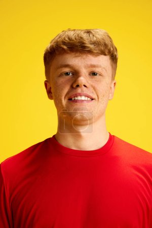 Téléchargez les photos : Portrait de jeune homme roux avec des taches de rousseur, portant un t-shirt rouge, souriant, regardant la caméra sur fond de studio jaune. Concept de mode de vie actif, de jeunesse, de passe-temps et d'émotions humaines - en image libre de droit