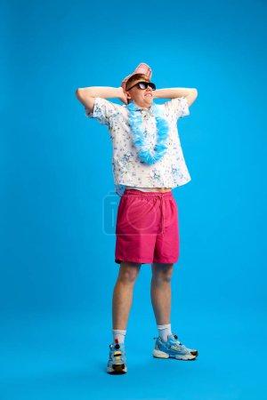 Foto de Retrato de cuerpo entero del joven con ropa elegante, camisa y camisas rosas, gafas de sol y sombrero de pie en pose relajante sobre fondo azul. Concepto de juventud, tiempo libre, vacaciones de verano - Imagen libre de derechos