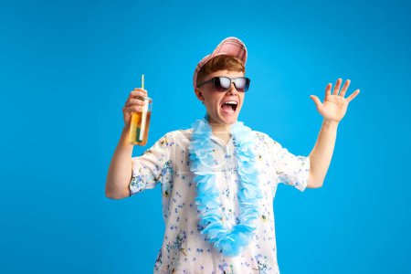 Photo for Emotional young man with flower necklace accessory, standing in sunglasses, holding lemonade and expressing excitement over blue studio background. Concept of emotions, youth, leisure time, vacation - Royalty Free Image