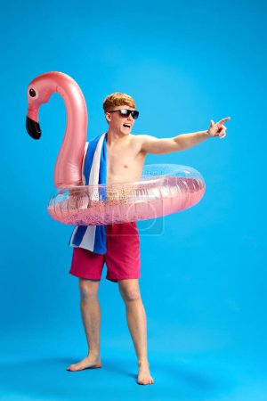 Téléchargez les photos : Jeune homme gai et détendu dans un cercle de natation profitant d'un voyage d'été, s'amusant sur fond de studio bleu. Concept d'émotions, de jeunesse, de temps libre, de vacances d'été - en image libre de droit