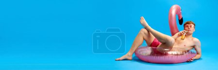 Téléchargez les photos : Jeune homme en short de bain couché sur un cercle de natation géant, sirotant de la limonade et se relaxant sur fond de studio bleu. Concept d'émotions, jeunesse, temps libre, vacances d'été. Bannière pour annonce - en image libre de droit