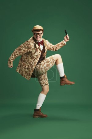Photo for Young smiling man in image of detective, wearing retro clothes, sneaking with magnifying glass against green studio background. Concept of imagination, retro style, fashion, youth, creativity - Royalty Free Image