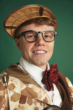 Téléchargez les photos : Portrait de jeune homme en lunettes, vêtu d'un costume vintage de détective, souriant, regardant la caméra sur fond de studio vert. Concept d'imagination, style rétro, mode, jeunesse, créativité - en image libre de droit