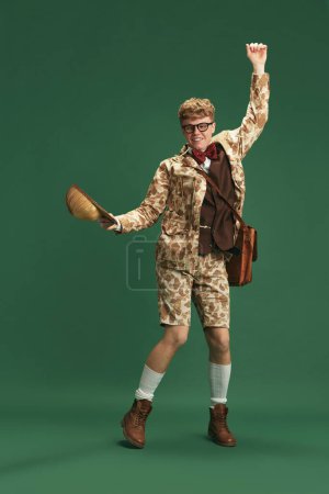 Photo for Full-length image of young guy in retro clothes in image of boy scout dancing, having fun against green studio background. Concept of imagination, retro style, fashion, youth, creativity - Royalty Free Image