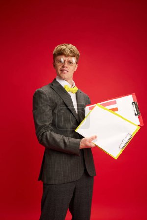 Photo for Young man, office worker in formal wear and glasses, holding many papers, projects against red studio background. Overworking. Concept of business, youth, human emotions, lifestyle - Royalty Free Image