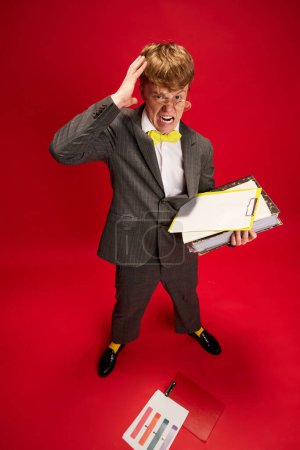 Photo for Deadlines. Young shocked man, employee in formal wear holding hand on head, looking on many business documents against red studio background. Concept of business, youth, human emotions, lifestyle - Royalty Free Image