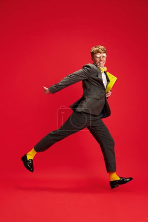 Photo for Full-length image of young man, employee holding documents and cheerfully running against red studio background. Motivation. Concept of business, youth, human emotions, lifestyle - Royalty Free Image