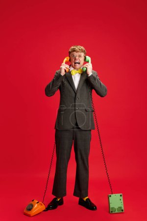 Photo for Full-length image of angry, emotional young man, employee, assistant standing with two phones against red studio background. Concept of business, youth, human emotions, lifestyle - Royalty Free Image