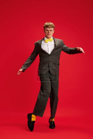 Foto de Retrato de cuerpo entero de un joven en ropa formal con calcetines amarillos brillantes bailando sobre fondo rojo del estudio. Concepto de negocio, juventud, emociones humanas, estilo de vida - Imagen libre de derechos
