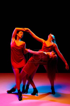 Téléchargez les photos : Jeunes belles femmes en robes rouges élégantes dansant, faisant des performances sur fond noir dans la lumière au néon. Concept de style de danse moderne, créativité et beauté, art, passe-temps - en image libre de droit