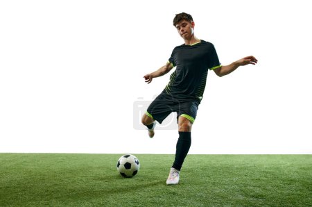 Foto de Joven, jugador de fútbol en movimiento durante el juego, golpeando pelota aislada sobre fondo blanco con suelo de hierba. Concepto de deporte, juego, competición, campeonato, estilo de vida activo - Imagen libre de derechos