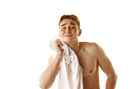 Foto de Hombre joven sin camisa de pie con toalla, lavando la cara aislada sobre fondo blanco. Rutina. Concepto de belleza masculina, cuidado de la piel, cosmetología y cosmética, salud - Imagen libre de derechos