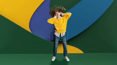 Photo for Youth culture in advertising for smart, casual clothing lines. Tech gadgets as part of modern lifestyle. Young, emotional woman listening to music in headphones and dancing over abstract background. - Royalty Free Image