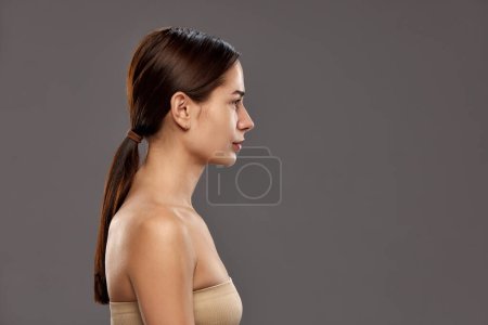 Photo for Profile view of beautiful young woman with ponytail, wearing strapless top against studio background. No makeup look. Concept of natural beauty, cosmetology and cosmetics, skin care - Royalty Free Image