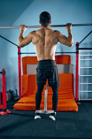 Photo for Young sportive man with muscular body, relief strong back training shirtless, doing pull ups exercises with weight in modern gym. Concept of active and healthy lifestyle, body care, fitness, sport - Royalty Free Image