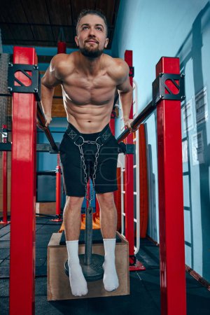 Téléchargez les photos : Musclé homme torse nu formation dans la salle de gym, faire des exercices de trempettes sur des barres parallèles avec ceinture pondérée autour de la taille pour la résistance. Concept de mode de vie actif et sain, soins du corps, fitness - en image libre de droit