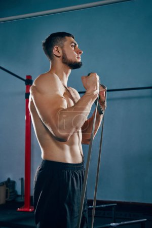 Photo for Young bearded man with muscular, relief strong body training in gym, doing exercises with fitness expanders. Concept of active and healthy lifestyle, body care, fitness, sport - Royalty Free Image