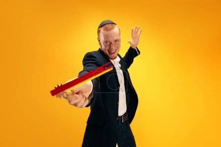 Photo for Portrait of emotional young Jewish man in yarmulke, black suit and with wooden noisemaker celebrating Purim against yellow studio background. Concept of holiday, Jewish traditions, history and culture - Royalty Free Image