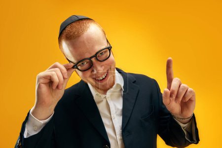 Téléchargez les photos : Portrait de jeune homme juif souriant en yarmulke, lunettes et costume levant le doigt sur fond de studio jaune. Votre attention. Concept de vacances Pourim, traditions juives, histoire et culture - en image libre de droit