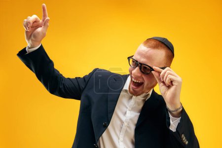 Téléchargez les photos : Portrait de jeune homme juif souriant et heureux en yarmulke, lunettes et costume levant le doigt sur fond de studio jaune. Concept de vacances Pourim, traditions juives, histoire et culture - en image libre de droit