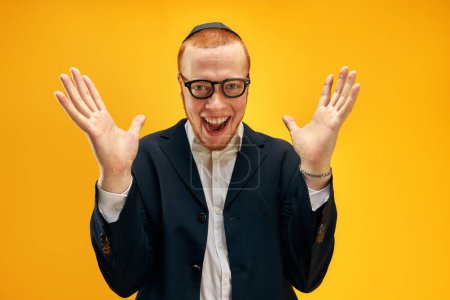 Téléchargez les photos : Portrait de jeune homme juif roux heureux dans des lunettes et yarmulke posant joyeusement sur fond de studio jaune. Concept de vacances Pourim, traditions juives, histoire et culture - en image libre de droit