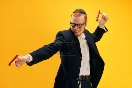 Téléchargez les photos : Jeune homme juif rousse émotionnelle dans des lunettes, yarmulke jouer avec bruyant en bois, gragger sur fond de studio jaune. Concept de vacances Pourim, traditions juives, histoire et culture - en image libre de droit
