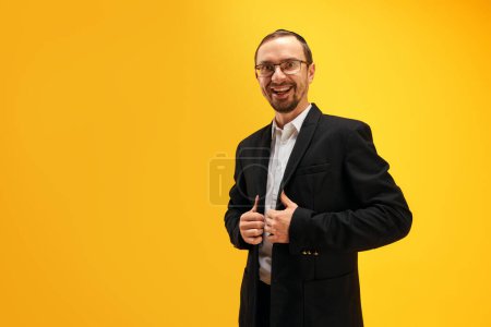 Téléchargez les photos : Portrait de l'homme juif émotionnel et excité en costume noir, yarmulke posant sur fond de studio jaune. Joyeuses fêtes. Concept de vacances Pourim, traditions juives, histoire et culture - en image libre de droit