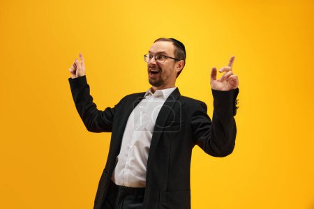 Photo for Portrait of emotional, excited Jewish man in black suit, yarmulke posing against yellow studio background. Happy holidays. Concept of Purim holiday, Jewish traditions, history and culture - Royalty Free Image