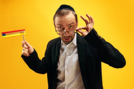 Photo for Portrait of man in his 30s wearing glasses, yarmulke and playing with noisemaker, gragger, expressing emotions over yellow studio background. Concept of Purim, Jewish traditions, history and culture - Royalty Free Image