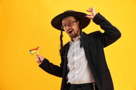 Téléchargez les photos : Joyeux homme juif au chapeau, avec des latéraux tenant bruyant, sur fond jaune. Fête. Concept de vacances Pourim, traditions juives, histoire et culture - en image libre de droit