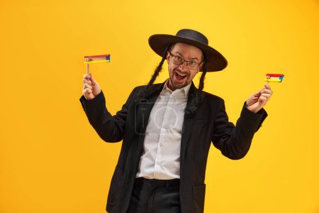 Photo for Cheerful Jewish man in hat, with sidelocks holding noisemaker, against yellow background. Celebration. Concept of Purim holiday, Jewish traditions, history and culture - Royalty Free Image