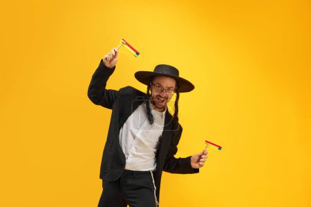 Photo for Cheerful Jewish man in hat, with sidelocks holding noisemaker, against yellow background. Celebration. Concept of Purim holiday, Jewish traditions, history and culture - Royalty Free Image