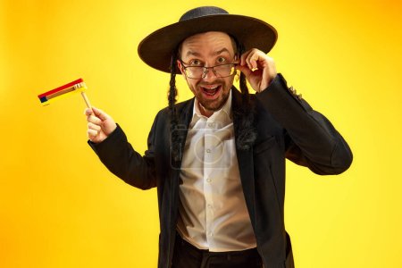 Téléchargez les photos : Jeune homme juif excité et émotionnel coiffé d'un chapeau, avec des latéraux, dans des lunettes debout avec un bruyant sur fond de studio jaune. Concept de vacances Pourim, traditions juives, histoire et culture - en image libre de droit