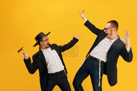 Two emotional Jewish man, friends in suits, yarmulke, with noisemaker having fun, celebrating against yellow studio background. Concept of Purim holiday, Jewish traditions, history and culture