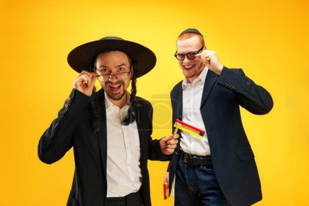Téléchargez les photos : Deux hommes juifs émotionnels, des amis en costume, yarmulke, avec bruyant s'amuser, célébrant sur fond de studio jaune. Concept de vacances Pourim, traditions juives, histoire et culture - en image libre de droit