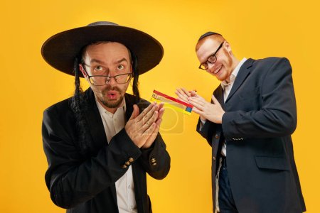 Photo for Two emotional Jewish man, friends in suits, yarmulke, with noisemaker having fun, celebrating against yellow studio background. Concept of Purim holiday, Jewish traditions, history and culture - Royalty Free Image
