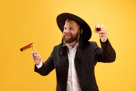 Photo for Cheerful young Jewish man in hat, with sidelocks holding noisemaker, against yellow background. Celebration. Concept of Purim holiday, Jewish traditions, history and culture - Royalty Free Image